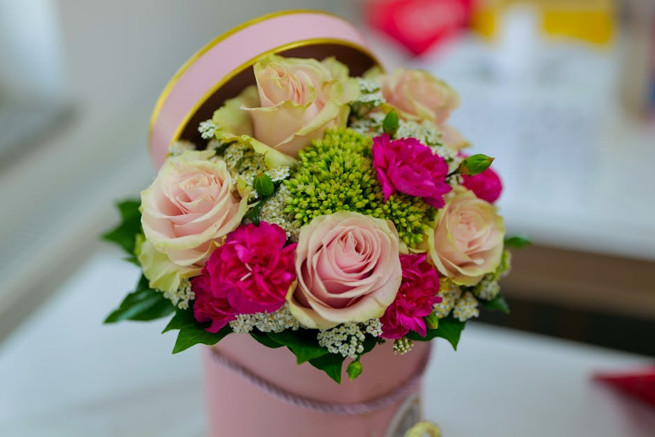 A round bouquet arrangement featuring pale pink roses complemented by vibrant magenta carnations and small white filler flowers, all set in a bed of lush green foliage. The flowers are neatly arranged