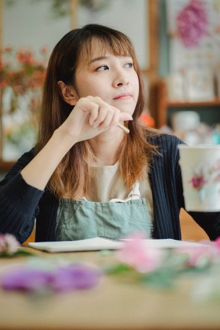 A close-up of a florist with shoulder-length brown hair, wearing a light beige top and a dark cardigan, seated at a table in a flower shop. She is holding a small flower with her right hand, inspectin