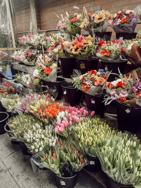 A display of various fresh flower bouquets at a flower shop, featuring a vibrant assortment including pink tulips, orange tulips, and white tulips arranged in black buckets. Behind the buckets, there 