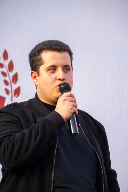 This image shows a young man with dark hair, wearing a black jacket and black shirt, holding a microphone close to his mouth as he appears to be speaking or singing. He is positioned against a plain l